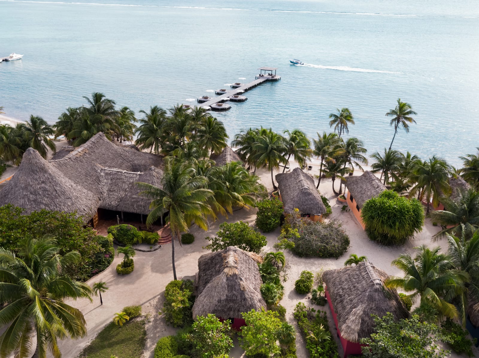 Aerial view of Matachica Resort & Spa — thatched-roof casitas among palms and a pier with umbrella-shaded lounge decks reaching into the turquoise Caribbean, Ambergris Caye, Belize