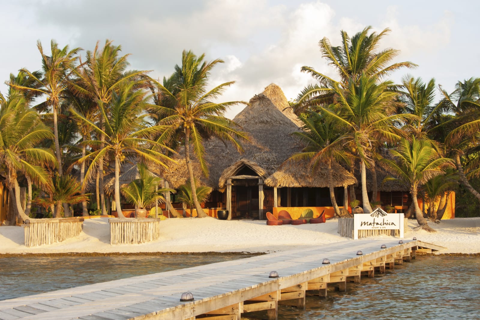 Matachica Resort & Spa seen from the water at golden hour, its thatched-roof beachfront pavilion framed by palms with the resort's sign at the end of the dock, Ambergris Caye, Belize