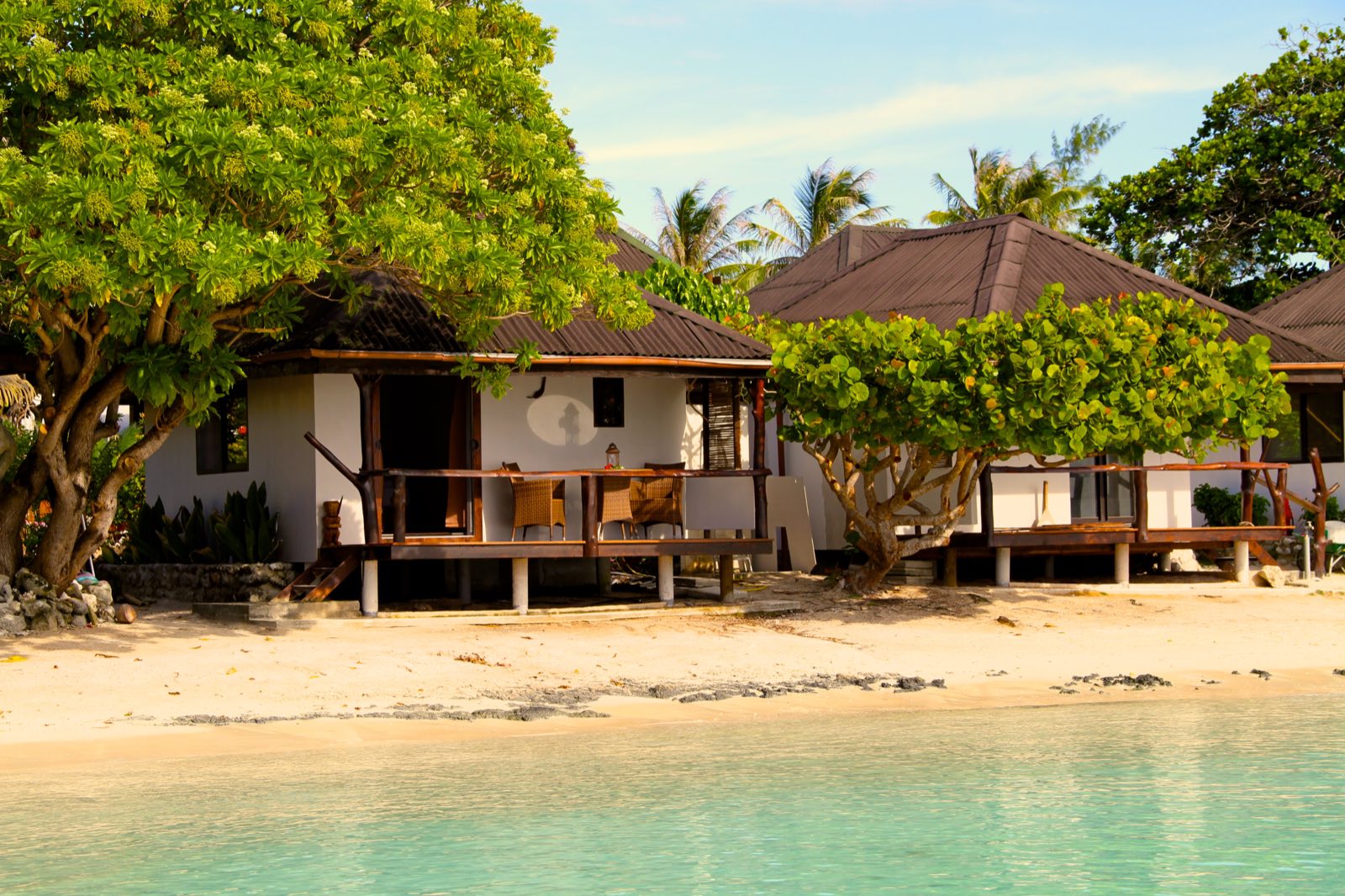 White beachfront bungalows with thatch-edged terraces at Havaiki Lodge, set on the sand between shade trees at the edge of the turquoise Fakarava lagoon
