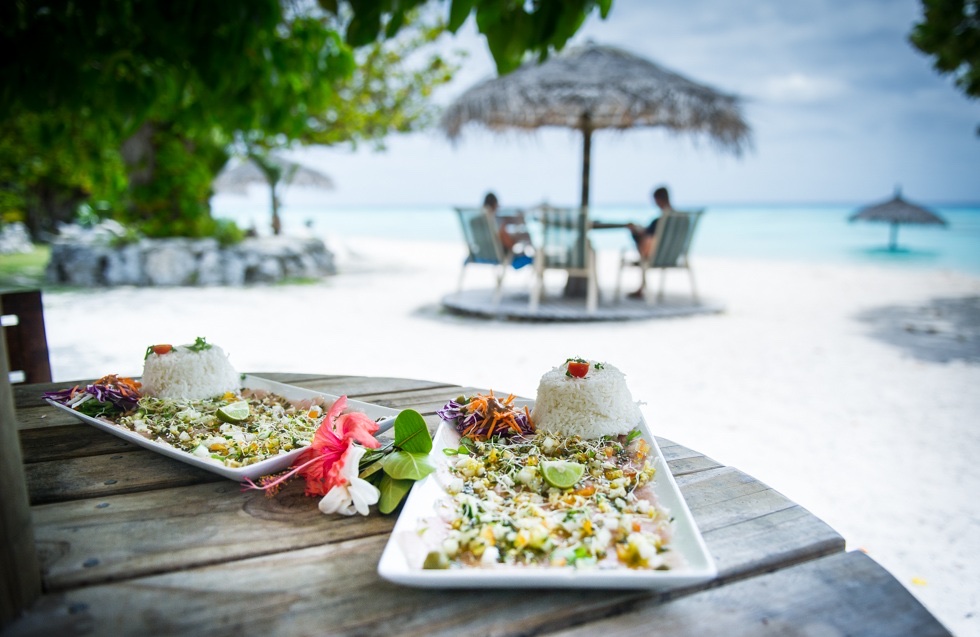 Plates of poisson cru and rice garnished with hibiscus flowers on a wooden beach table at Havaiki Lodge, with a thatched-umbrella dining spot on the white sand behind