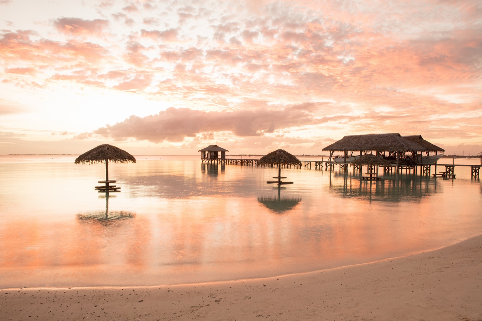 Thatched-umbrella tables standing in the shallow lagoon at Havaiki Lodge under a pink and gold Fakarava sunset, with the over-water pearl farm on its jetty behind them