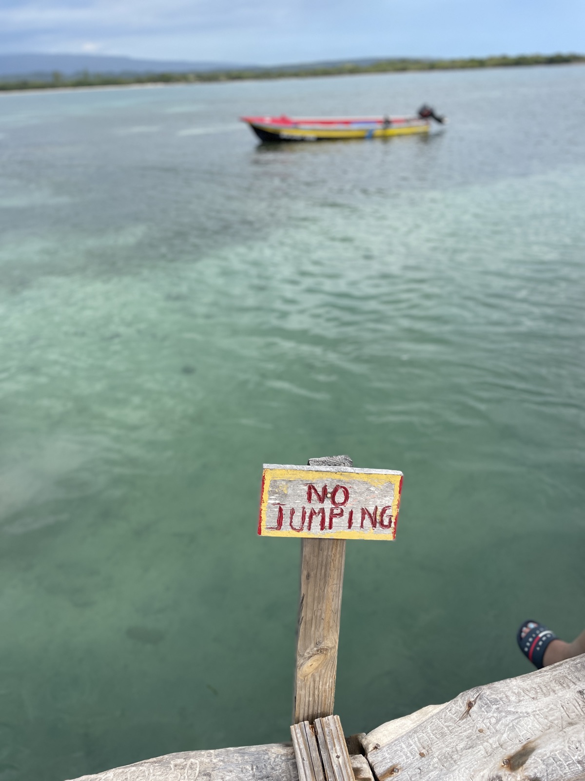 A hand-painted 'No Jumping' sign at Floyd's Pelican Bar with a colorful boat and clear shallow water behind it