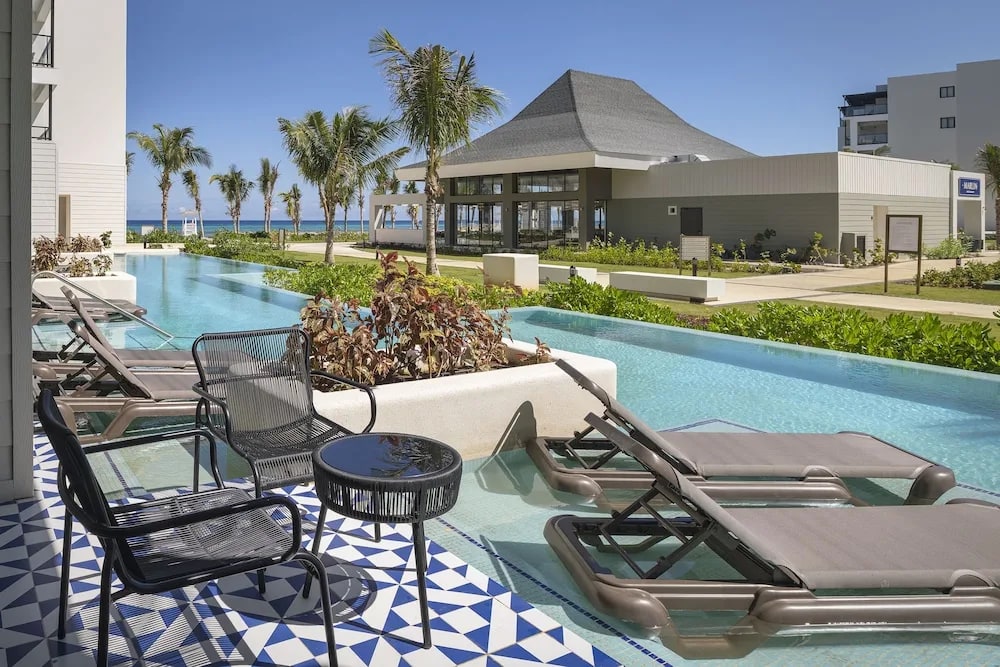 Swim-up suite at Ocean Eden Bay, loungers set into the shallow edge of the channel pool on patterned blue tile, the water running on past the neighboring terraces toward the sea